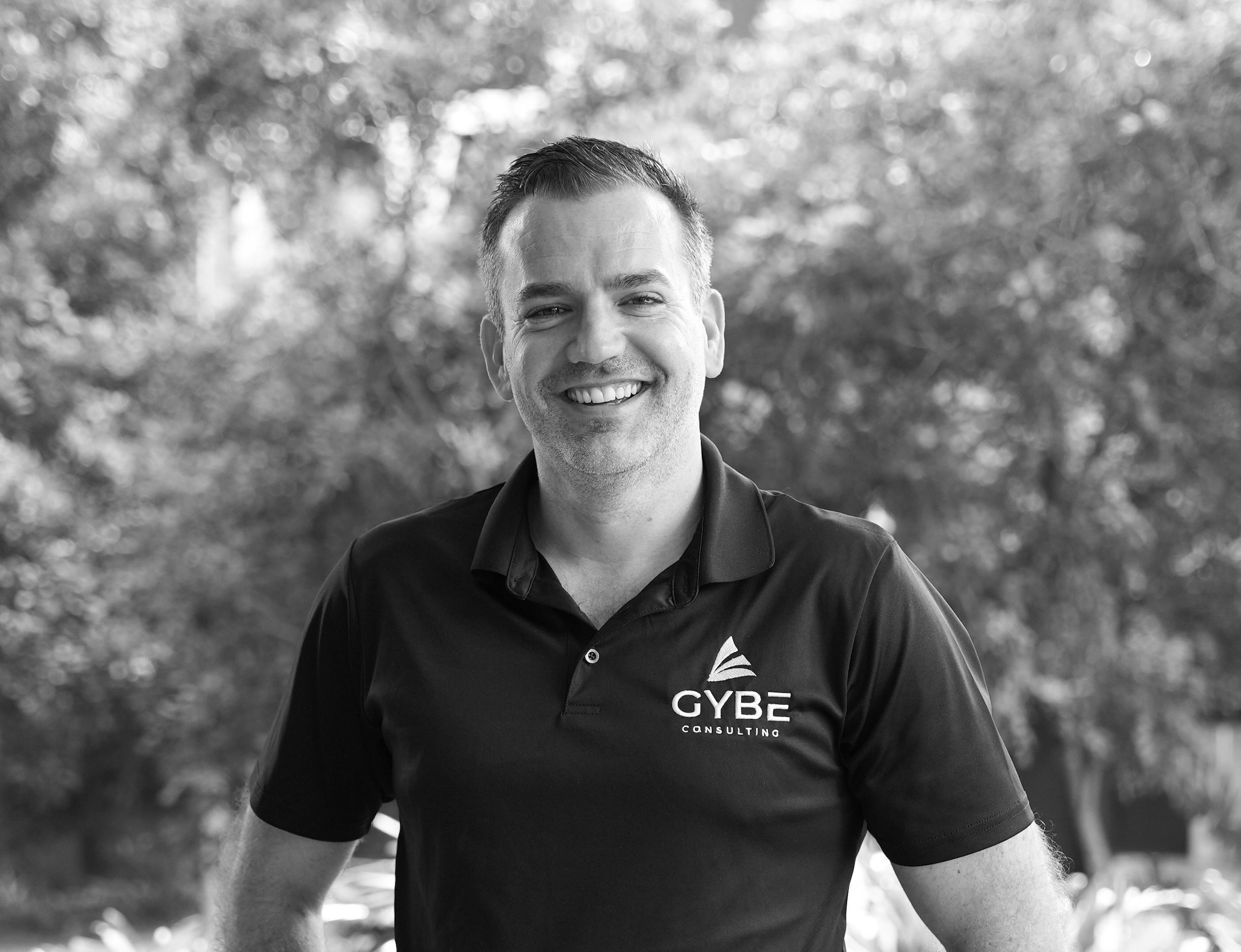 Smiling man wearing a black polo shirt with GYBE Consulting logo standing outdoors with blurred trees in the background.